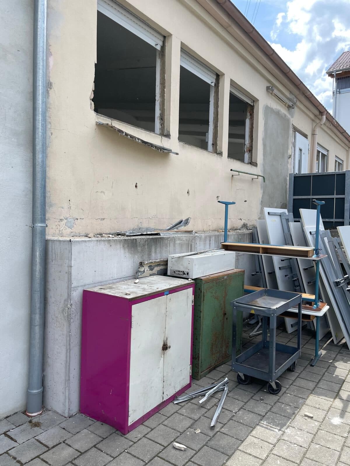 Außenwand des alten Gebäudes mit ausgebauten Fenstern, davor alte Schränke, Regale und Tafeln