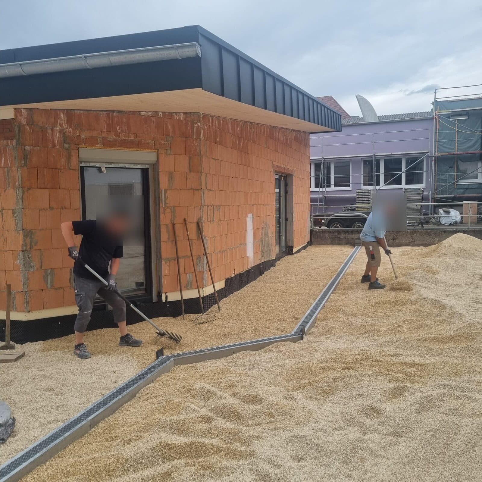 Zwei Helfer verteilen Kies um den Neubau, dazwischen eine neue Entwässerungsrinne