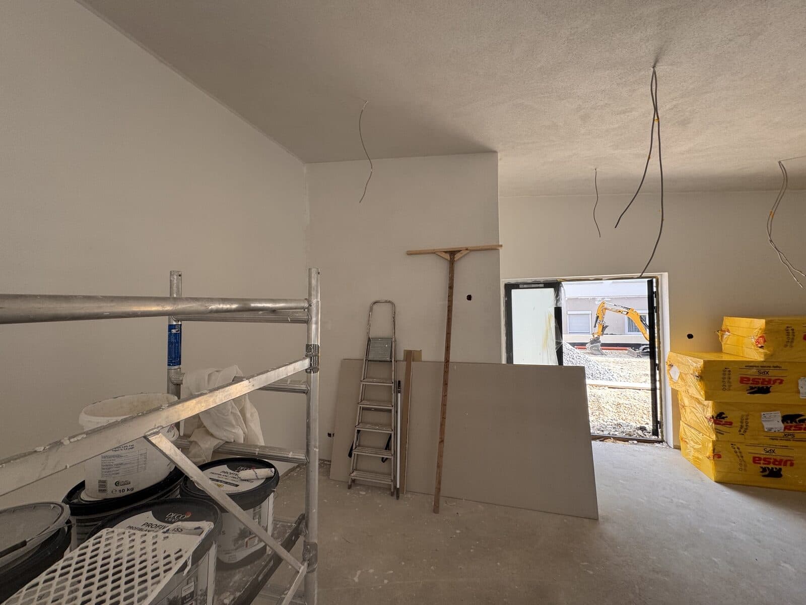 Die Küche im Rohbau: Gerüst, Farbeimer und Gipskartonplatten, offene Kabel, Tür nach draußen
